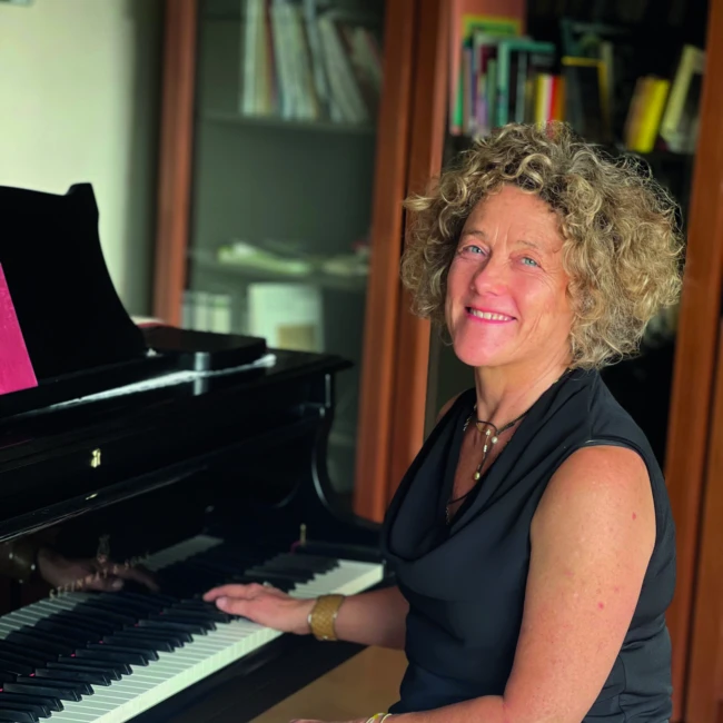 <think> Got it, let's create the alt text. First, identify what's visible: a woman with curly hair sitting at a grand piano…